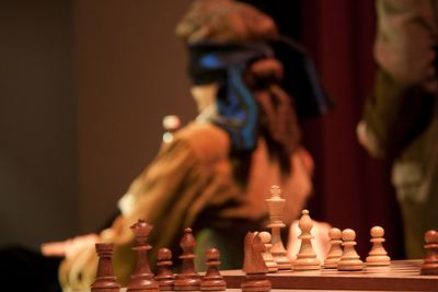 Holzfiguren mit weißem König und Bauern im Vordergrund, unscharf dahinter der Darsteller mit blauer Augenbinde