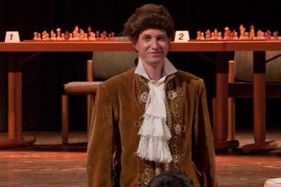 Der Darsteller mit Perücke lächelt ohne Augenbinde, hinter ihm die Spieltafel mit den Brettern 1 und 2