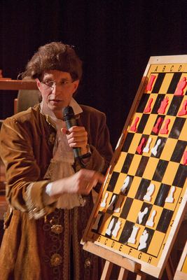 Der Darsteller im Samtrock zeigt mit Mikrofon in der Hand auf die weißen Figuren am gelb-schwarzen Demobrett