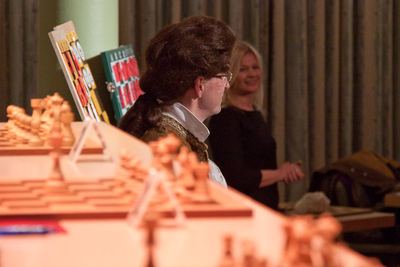 Der Darsteller mit Zopfperücke im Profil hinter den Schachbrettern, im Hintergrund lächelt die FIDE-Meisterin