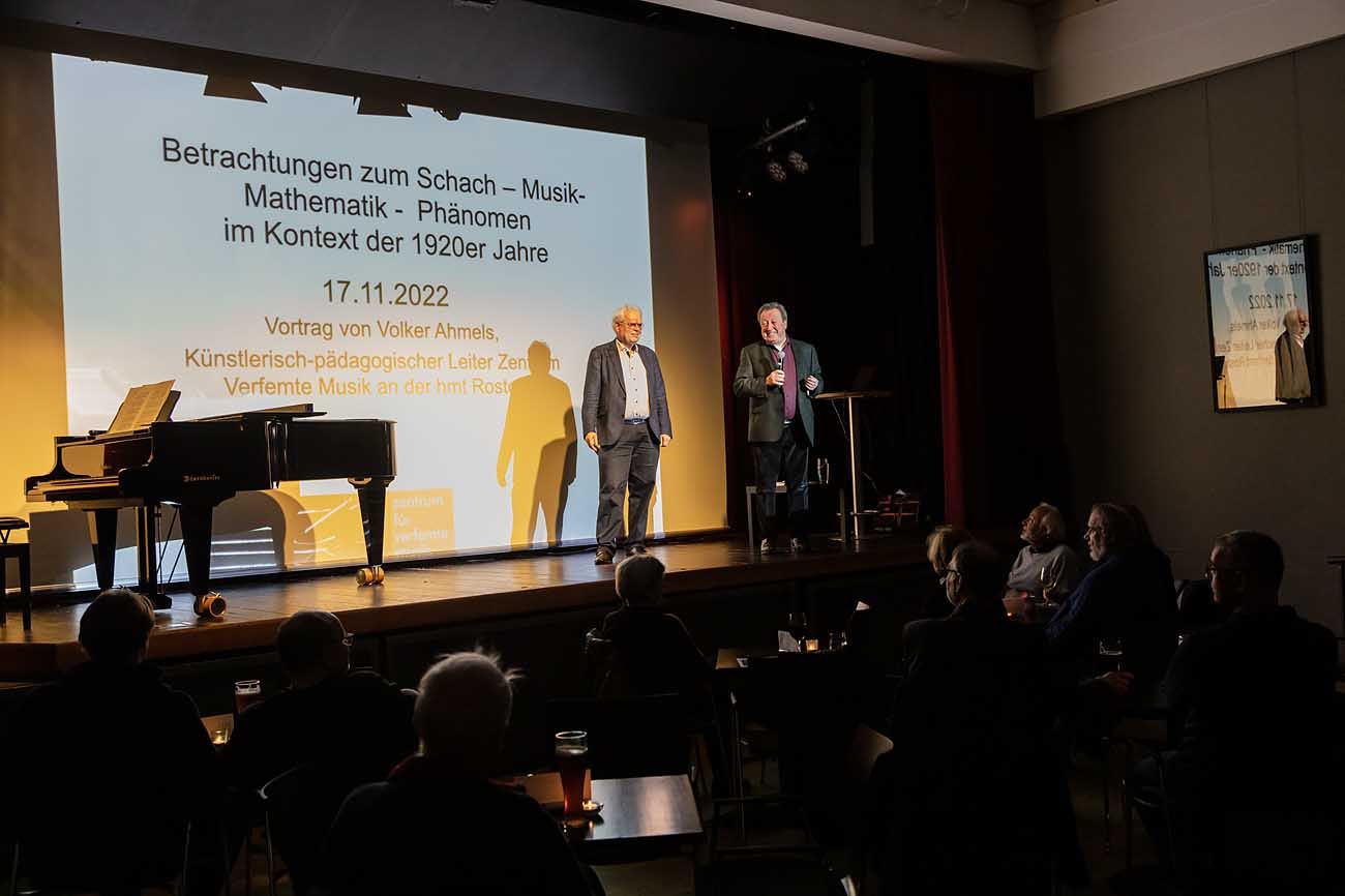 Moderator mit Mikrofon begrüßt den Referenten auf der Bühne des Alten Kinos, dahinter die Titelfolie zum Schach-Musik-Mathematik-Phänomen der 1920er Jahre