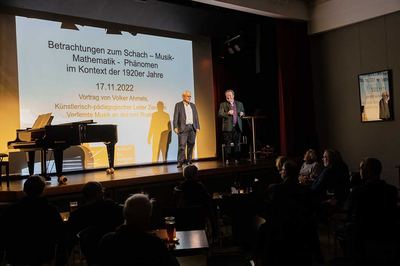 Moderator mit Mikrofon begrüßt den Referenten auf der Bühne des Alten Kinos, dahinter die Titelfolie zum Schach-Musik-Mathematik-Phänomen der 1920er Jahre