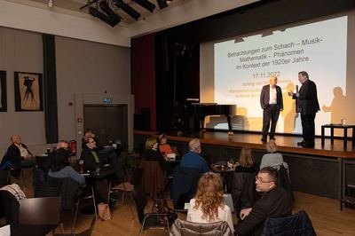 Referent und Moderator im Gespräch auf der Bühne neben einem Flügel, im Saal Zuhörer an Bistrotischen