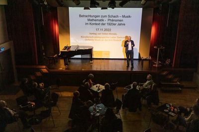 Blick von oben: Der Referent spricht vor der Titelprojektion seines Vortrags, links ein Flügel, davor Publikum an Tischen
