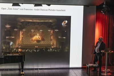 Referent blickt auf die Leinwand mit einem Ausschnitt einer Opernaufführung in einem barocken Theatersaal