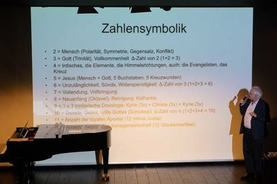 Referent mit Mikrofon vor einer Folie zur Zahlensymbolik in der Musik, links ein Flügel