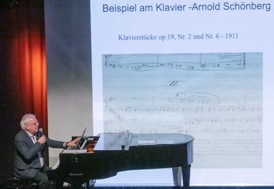 Referent spielt am Flügel ein Musikbeispiel, auf der Leinwand Notenausschnitte von Klavierstücken aus dem Jahr 1911