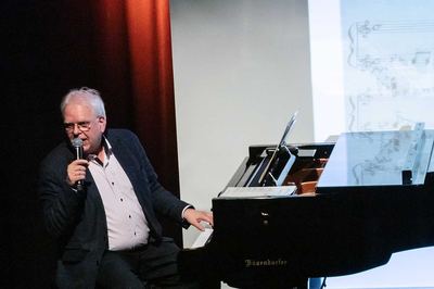 Referent am Flügel erläutert mit Mikrofon in der Hand ein Klavierstück, im Hintergrund projizierte Noten