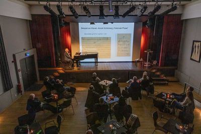 Blick über den Saal mit Zuhörern an Tischen auf die Bühne, wo der Referent am Flügel vor projizierten Notenblättern sitzt