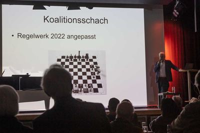 Folie „Koalitionsschach“ mit Foto eines Schachbretts für mehrere Spieler, der Referent erläutert die angepassten Regeln