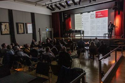 Saal im Alten Kino mit Publikum an Tischen, auf der Leinwand ein Plakat mit Schachdiagrammen und Zugmustern
