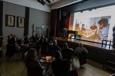 Publikum schaut auf eine Projektion, die vier Männer beim Koalitionsschach an einem Tisch zeigt