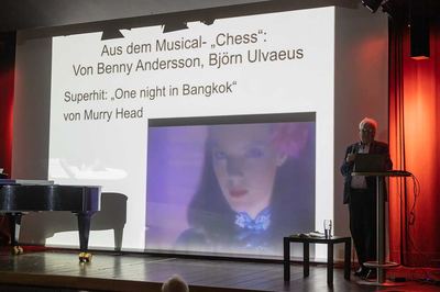 Referent am Stehpult vor einer Folie zum Musical „Chess“ mit Standbild aus einem Musikvideo