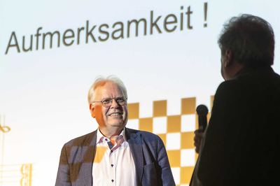 Lächelnder Referent nach dem Vortrag vor der Schlussfolie mit Schachbrettmuster, rechts der Moderator mit Mikrofon