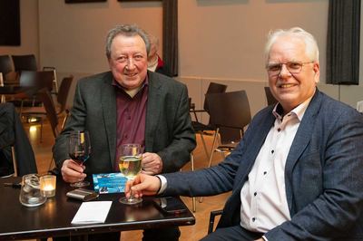 Zwei Männer mit Wein- und Weißweinglas sitzen lächelnd an einem Tisch im Saal nach dem Vortrag