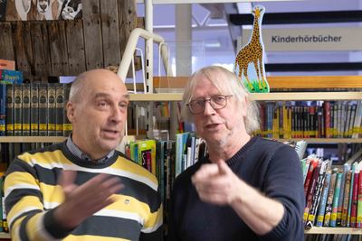 Zwei Männer gestikulieren im Gespräch vor Bücherregalen der Kinderabteilung, darüber eine bemalte Holzgiraffe