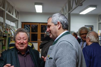 Ein Mann im grauen Trachtenjanker im Gespräch mit einem lachenden Gast im Sakko, im Hintergrund weitere Besucher