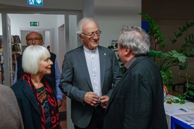 Begrüßung im Eingangsbereich der Bücherei: eine weißhaarige Dame mit rotem Schal und zwei Herren im Gespräch neben einer Grünpflanze