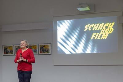 Lachende Moderatorin in Rot vor der Leinwand mit dem Ausstellungstitel „Schach und Film“, daneben mexikanische Aushangplakate