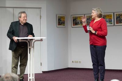 Moderatorin in rotem Pullover begrüßt den Redner am weißen Stehtisch, an der Wand Plakate zum Film „Jaque a la locura“