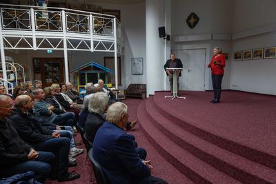 Eine Rednerin in roter Bluse spricht auf dem Podest mit rotem Teppich, daneben ein Mann am Stehtisch, links das sitzende Publikum