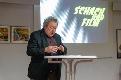 Redner am weißen Stehtisch vor der Leinwand mit dem leuchtenden Ausstellungstitel „Schach und Film“, links gerahmte Filmplakate