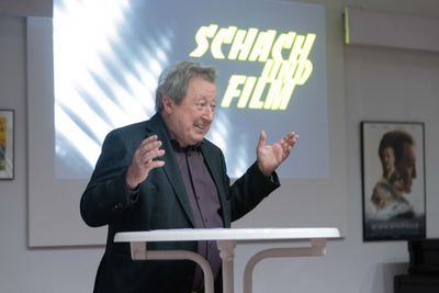 Redner am Stehtisch breitet bei der Eröffnungsrede die Arme aus, hinter ihm die Leinwand „Schach und Film“ und ein Schachnovelle-Plakat