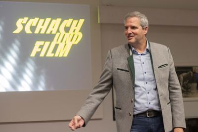 Redner im grauen Trachtenjanker spricht lächelnd vor der Projektion mit dem gelben Schriftzug „Schach und Film“
