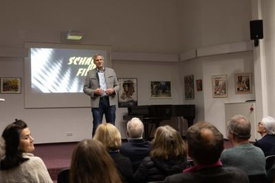 Redner im Trachtenjanker vor Publikum, an den Wänden Filmplakate wie „Schachnovelle“ und „Scacco alla follia“, daneben ein Flügel