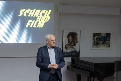 Redner im dunklen Sakko steht vor der Leinwand, rechts die Plakate zu „Schachnovelle“ und „Scacco alla follia“ über einem Flügel