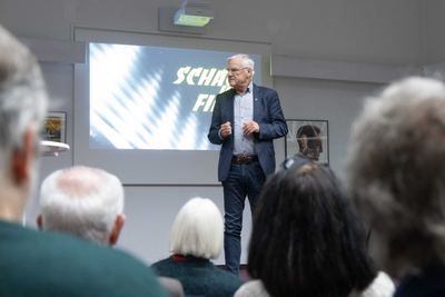Redner im blauen Sakko spricht vor der Projektion „Schach und Film“, im Vordergrund unscharf die Köpfe der Zuhörer