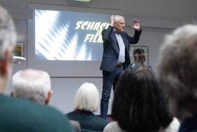Redner hebt beim Vortrag beide Hände, hinter ihm die Leinwand mit dem Ausstellungstitel, vorn die Hinterköpfe des Publikums