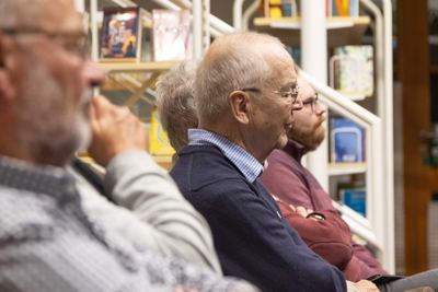 Zuhörer im Profil verfolgen konzentriert den Vortrag, im Hintergrund weiße Regale mit Büchern