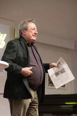 Redner hält bei seinem Vortrag eine aufgeschlagene englischsprachige Zeitungsseite mit einem Schachartikel hoch