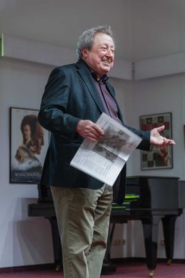 Lachender Redner mit aufgeschlagener Zeitung vor einem schwarzen Flügel, dahinter das Filmplakat „Schachnovelle“