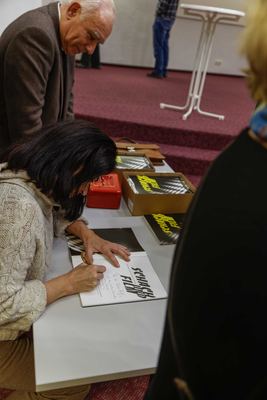 Eine Frau schreibt eine Widmung in den Katalog „Schach und Film“, daneben Katalogkartons und eine rote Geldkassette