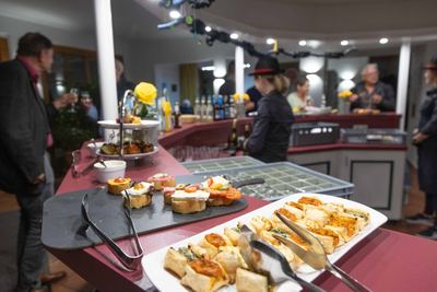 Fingerfood-Buffet mit belegten Baguettescheiben und Blätterteigtaschen, dahinter Gäste und Servicekräfte an der Bar