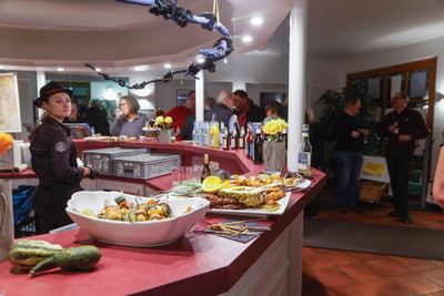 Buffet an der roten Theke mit Gemüsespießen und Fleischplatte, daneben eine Servicekraft mit schwarzem Hut, hinten plaudernde Gäste