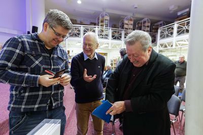 Drei Männer vor dem Vortrag im Gespräch, zwei betrachten lächelnd kleine Mitbringsel, dahinter die Galerie der Stadtbücherei