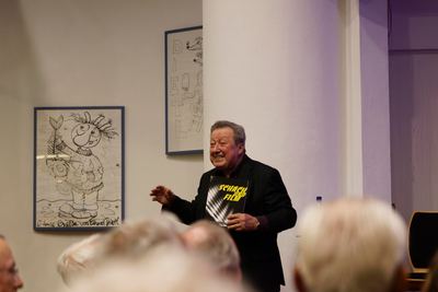Lachender Moderator im dunklen Sakko hält den Ausstellungskatalog „Schach und Film“ hoch, im Vordergrund unscharfe Zuhörerköpfe