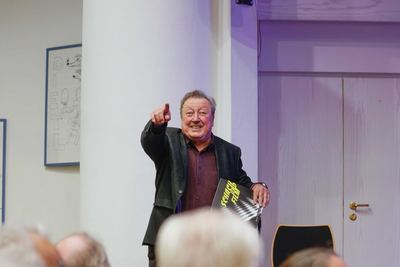 Der Moderator zeigt lachend ins Publikum, unter dem Arm den Katalog „Schach und Film“, vor einer weißen Säule