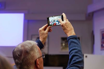 Ein Zuhörer hält sein Smartphone hoch und fotografiert das Publikum, im Hintergrund die beleuchtete Beamerleinwand