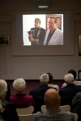 Filmszene vom Lebendschach auf der Leinwand: kostümierter König mit Krone und Zepter neben einem lachenden Mann, davor Zuhörer
