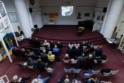 Blick von der Galerie auf den Vortragsraum: Zuhörer in Stuhlreihen schauen Lebendschach-Film, vorne Demobrett, Flügel und Leinwand