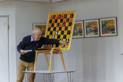 Der Großmeister beugt sich zum Demonstrationsbrett und zieht eine weiße Figur, an der Wand gerahmte Kinoplakate mit Schachmotiv