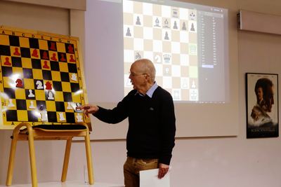 Der Großmeister deutet auf eine Mittelspielstellung am Demonstrationsbrett, hinter ihm die gleiche Stellung groß per Beamer projiziert