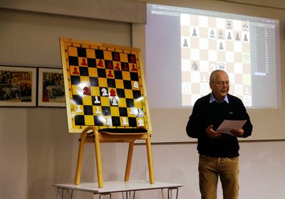 Der vortragende Großmeister mit Notizblatt neben dem Demonstrationsbrett mit Mittelspielstellung und der projizierten Partie