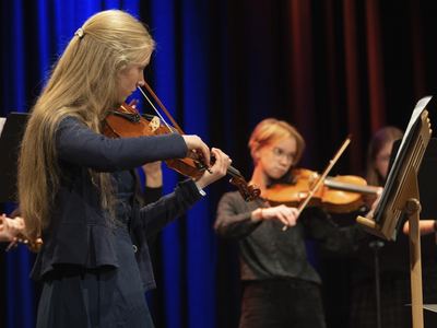 Junge Geigerinnen des Musikschul-Orchesters spielen auf der Bühne vor blau und rot beleuchtetem Vorhang, vorn ein hölzernes Notenpult