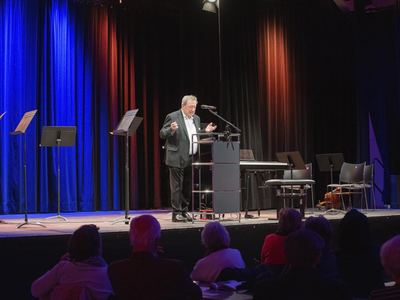 Redner mit Fliege am Pult auf der Bühne, daneben Notenpulte, Digitalpiano und ein abgelegtes Cello, vorn Zuschauer von hinten