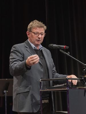 Grußwort-Redner in grauem Sakko gestikuliert am Mikrofon des Rednerpults vor dunklem Bühnenvorhang